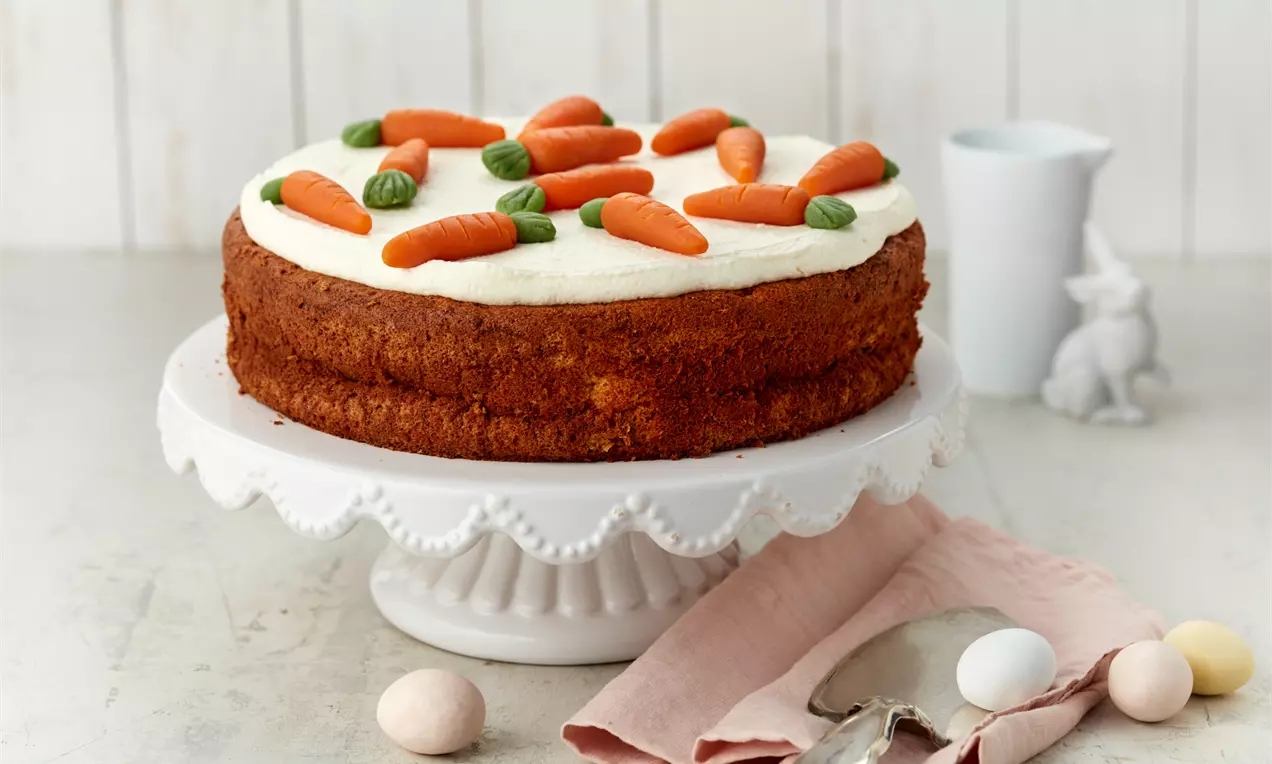 Karottenkuchen mit cremeweißer Glasur, dekoriert mit orangefarbenen Karotten aus Marzipan und grünen Blättern auf einer weißen Tortenplatte