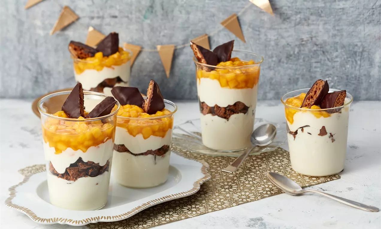 Schichtdessert im Glas mit weißer Mascarponecreme, braunen dunklen Keksstücken, orange-gelber Fruchtmasse und Dekor aus Schokoladenspitzen