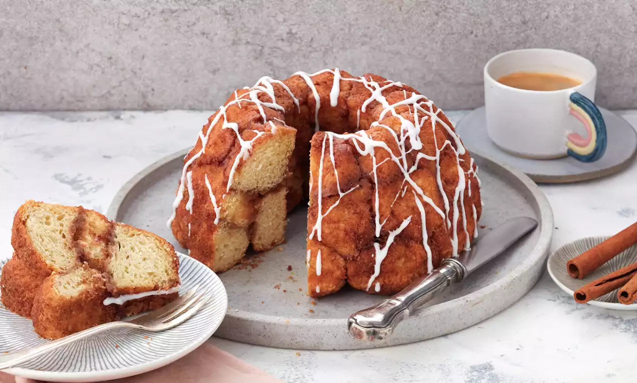 Monkey Bread mit karamellbrauner, zuckerbestreuter Oberfläche in Gugelhupfform, teilweise angeschnitten auf grauem Teller, weißer Zuckerguss