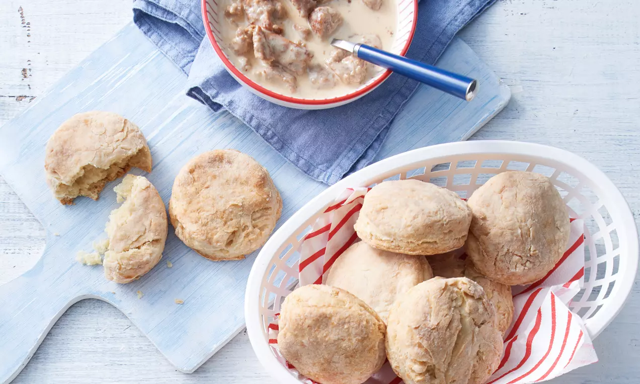 Goldbraune, leicht bröselige Biscuits in einem weißen Korb mit gestreiftem Tuch neben Schüssel mit sahniger Soße und Fleischstücken