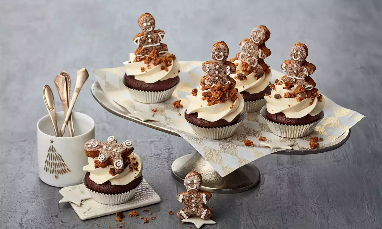 Dunkelbraune Lebkuchen-Cupcakes mit weißer Cremehaube, knusprigen Streuseln und Lebkuchenfiguren auf silberner Tortenplatte mit Stoffserviette