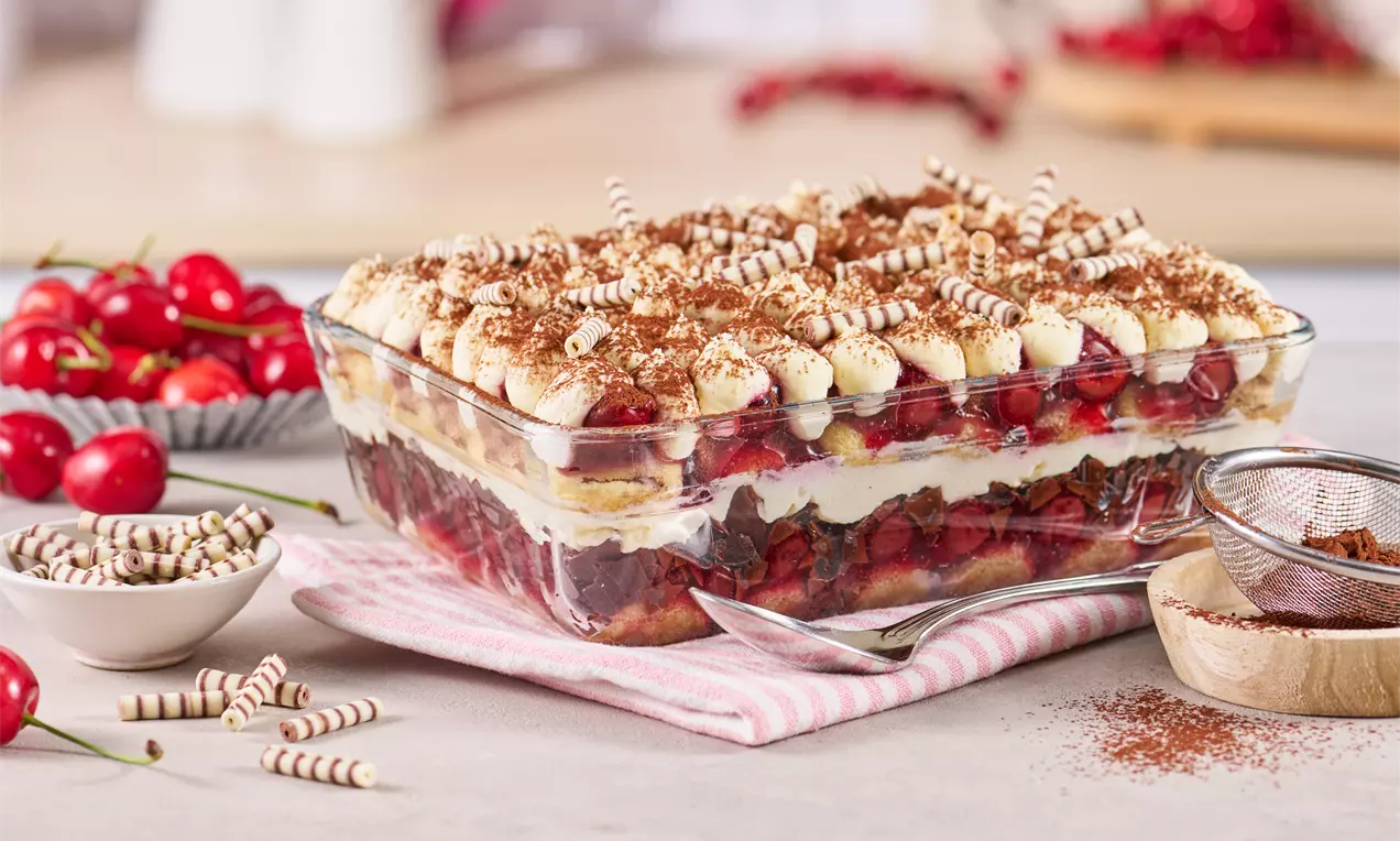 Ein mehrschichtiges Kirsch-Tiramisu mit Sahnetupfen, Kakao und Schokoröllchen in einer gläsernen Form auf gestreiftem Stoff