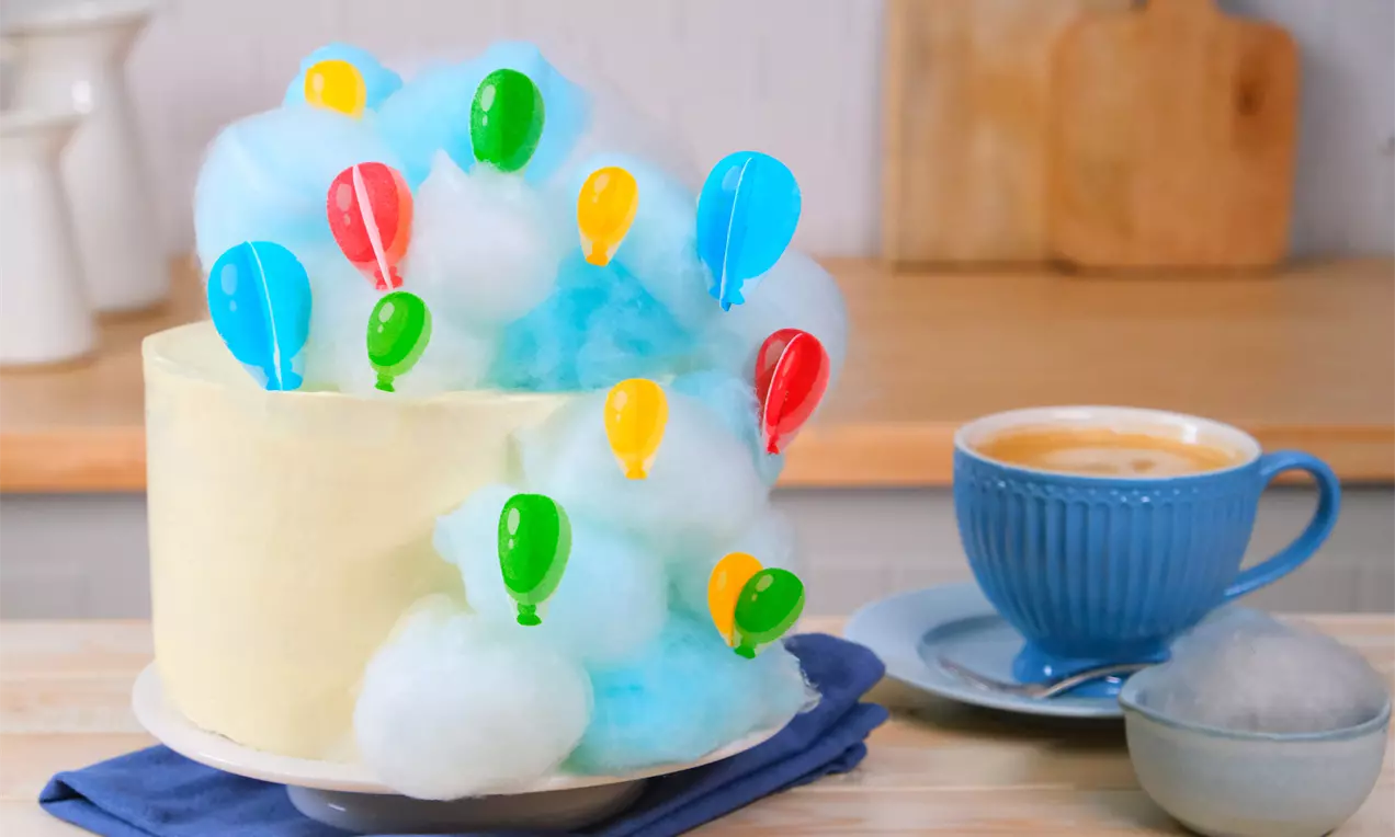 Kleine Zuckerwatte Torte - süß dekoriert mit weißer und blauer Zuckerwatte als Wolken um eine hohe Torte. In den Zuckerwatte Wolken stecken bunte Esspapier Luftballons.