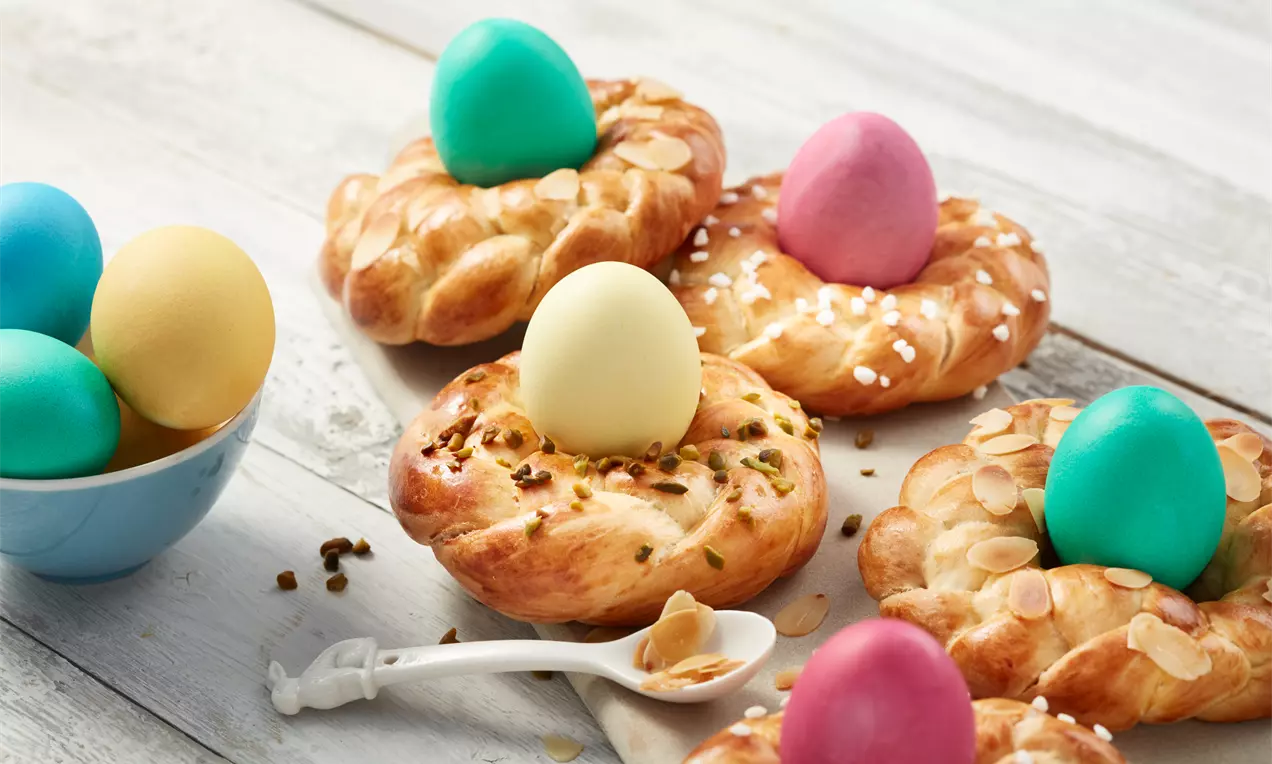 Goldbraune, geflochtene Osterkränzchen mit Mandeln, Pistazien und Zucker, dekoriert mit farbigen Ostereiern, arrangiert auf hellem Holz