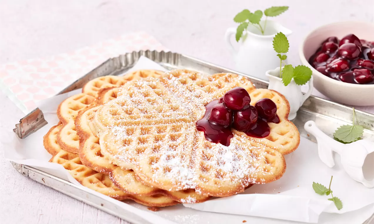 Herzförmige, goldbraune Waffeln mit Puderzucker bestäubt, dekoriert mit glänzend roten Kirschen auf einem Tablett mit heller Serviette