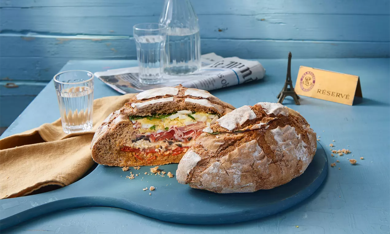 Rundes, goldbraunes Brot mit Mehl bestäubt, mittig aufgeschnitten, gefüllt mit bunten Schichten Gemüse und Belag auf blauem Holzbrett
