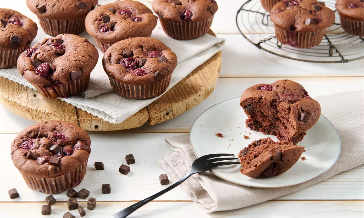 Saftige Schoko-Kirsch-Muffins mit Schokoladenstückchen, auf Holzbrett und Kuchengitter angerichtet, daneben ein angeschnittener Muffin auf Teller