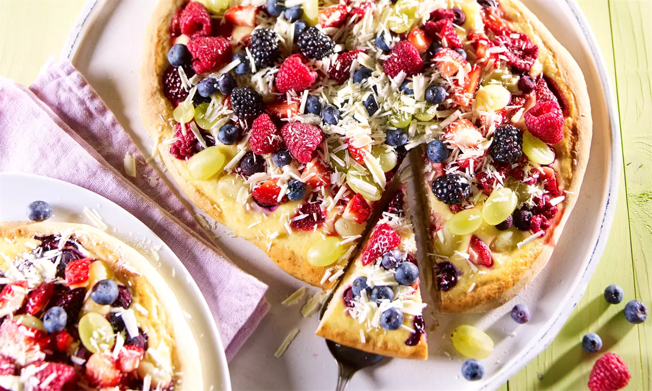 Helle Dessert-Pizza mit bunten Früchten wie Beeren und Trauben, geraspelter weißer Schokolade und einem herausgehobenen Stück auf hellen Tellern