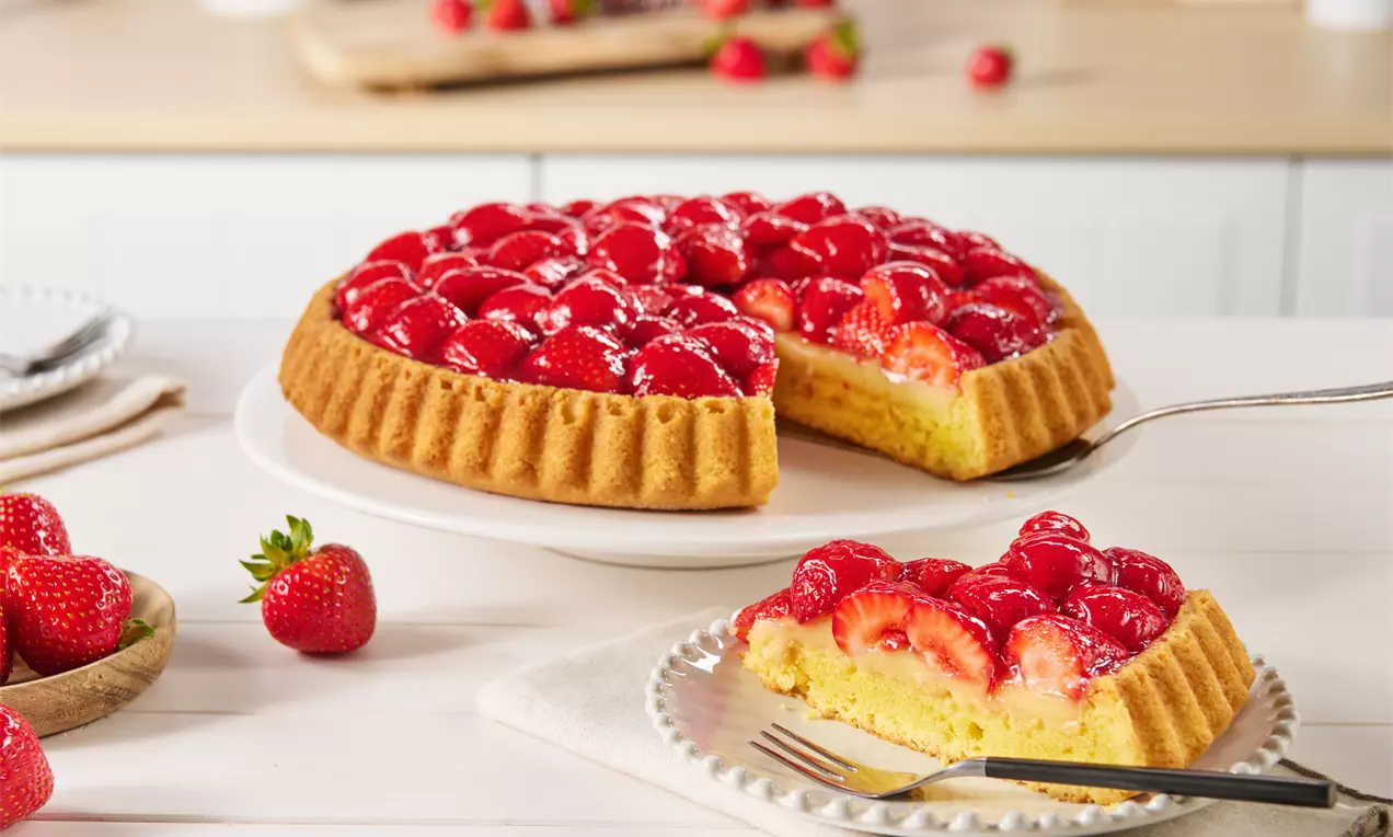 Veganer Erdbeerkuchen mit glänzender roter Erdbeerschicht auf hellem Bisquitteig, angeschnitten auf weißem Teller neben frischen Erdbeeren