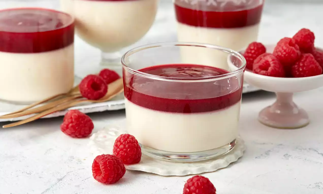 Weiße Joghurt-Panna-Cotta in einem klaren Glas, bedeckt von samtig roter Himbeersauce, arrangiert mit frischen Himbeeren auf hellem Hintergrund