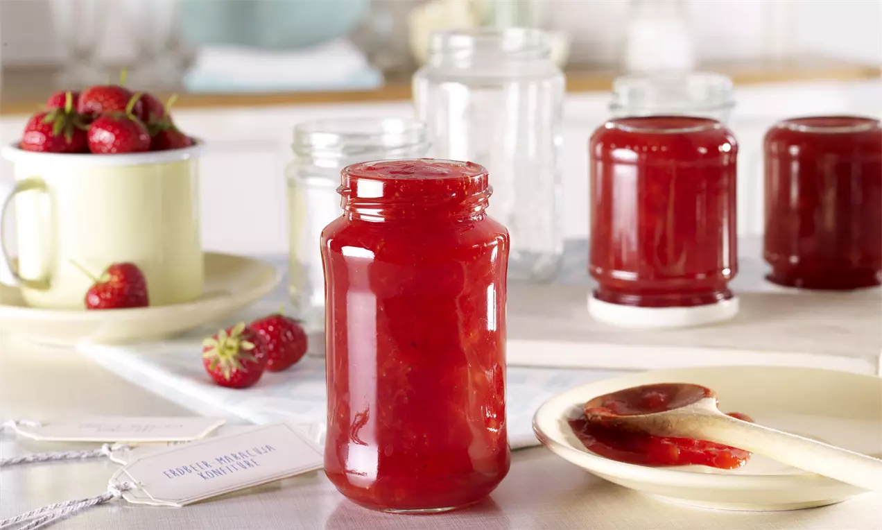 Leuchtend rote Erdbeer-Maracuja-Konfitüre in einem klaren Schraubglas, umgeben von frischen Erdbeeren und einem Holzlöffel mit Konfitüre