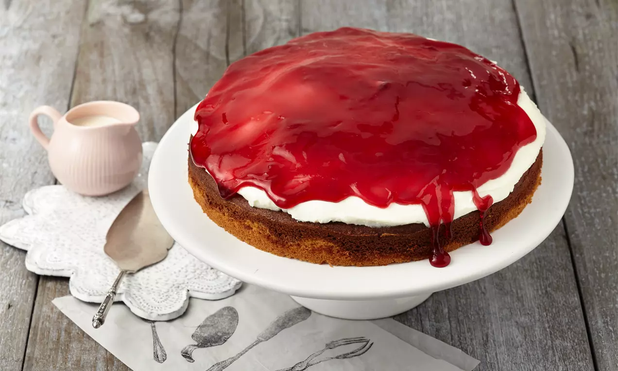Rotkäppchen-Kuchen mit goldenem Biskuitboden, weißer Quarkcreme und glänzender roter Kirschschicht auf einer weißen Tortenplatte.