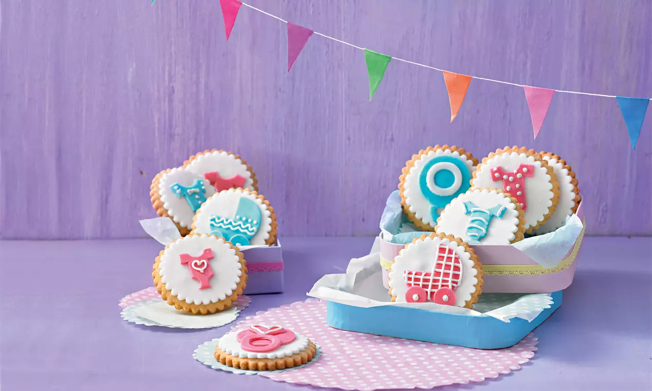 Runde Babykekse mit weißer Glasur, bunten Motiven wie Kinderwagen und Schnuller, teils in einer Box auf rosa Papier drapiert