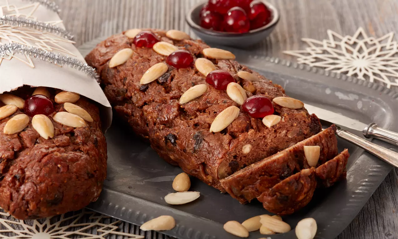 Früchtebrot in dunkler, rustikaler Optik mit glänzenden roten Kirschen und hellen Mandeln auf grauem Tablett mit winterlicher Dekoration