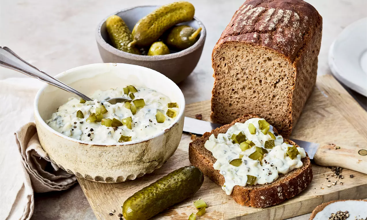 Gewürzgurken-Dip mit cremiger weißer Joghurtsauce, grünen Gewürzgurkenstückchen und Dill in weißer Schale, dazu knuspriges Bauernbrot