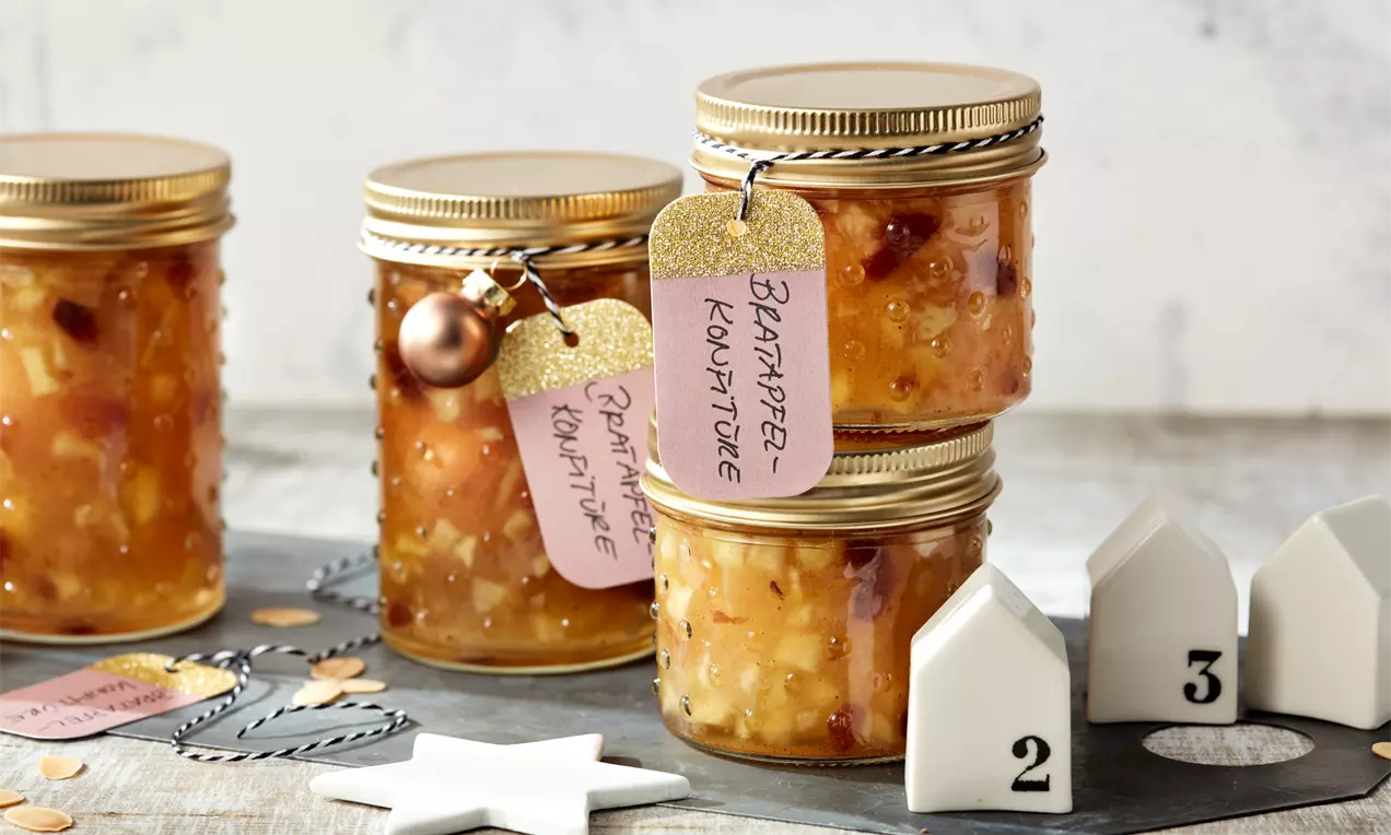 Bratapfelmarmelade im Glas mit goldbrauner, stückiger Konsistenz, Mandelsplittern und Zimtstangen neben einem Holzlöffel