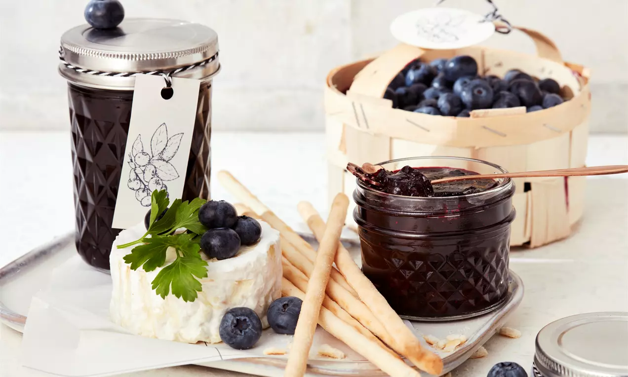Gläser mit dunkelvioletter Blaubeermarmelade, daneben ein Blauschimmelkäse, Grissini, frische Blaubeeren und ein Holzkorb voller Blaubeeren