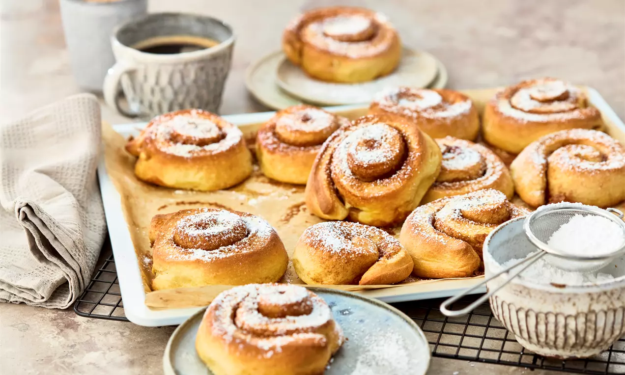 Goldbraune schwedische Zimtschnecken mit Zimtspiralen und Puderzucker auf einem weißen Backblech, begleitet von Tassen und Sieb