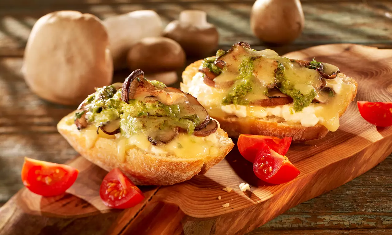 Goldbraune Röstbrote mit geschmolzenem Käse, Champignons und grünem Pesto auf einem Holzbrett, umgeben von frischen Kirschtomaten