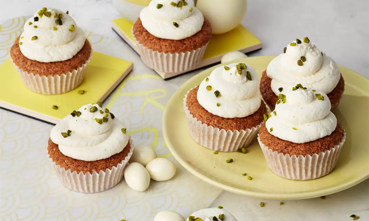Carrot-Cake-Muffins mit lockerer weißer Cremehaube und gehackten Pistazien bestreut auf gelbem Teller und hellem Untergrund arrangiert