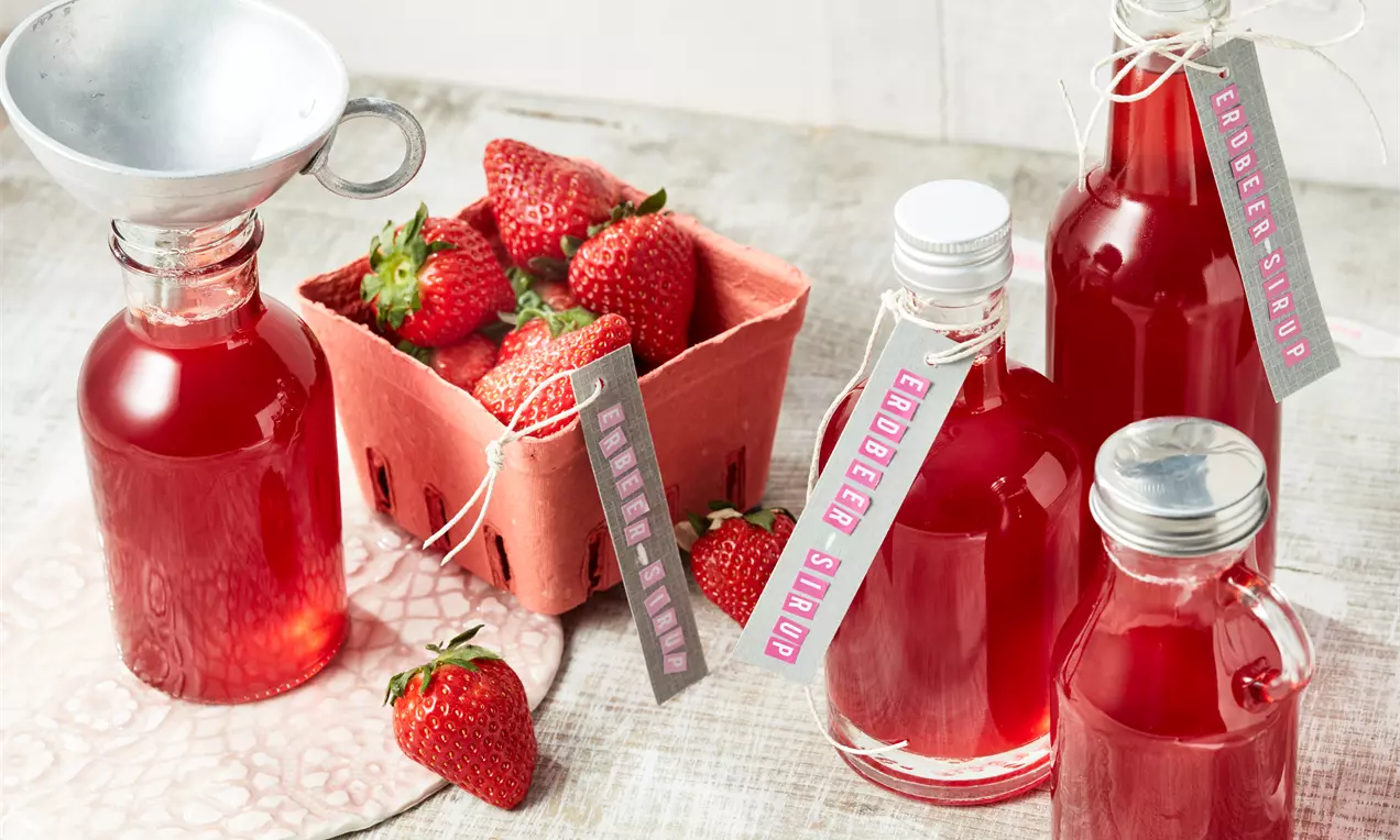 Rote Glasflaschen mit glänzendem Erdbeersirup, dekorative Etiketten, frische Erdbeeren in roter Pappschale und ein Metalltrichter daneben