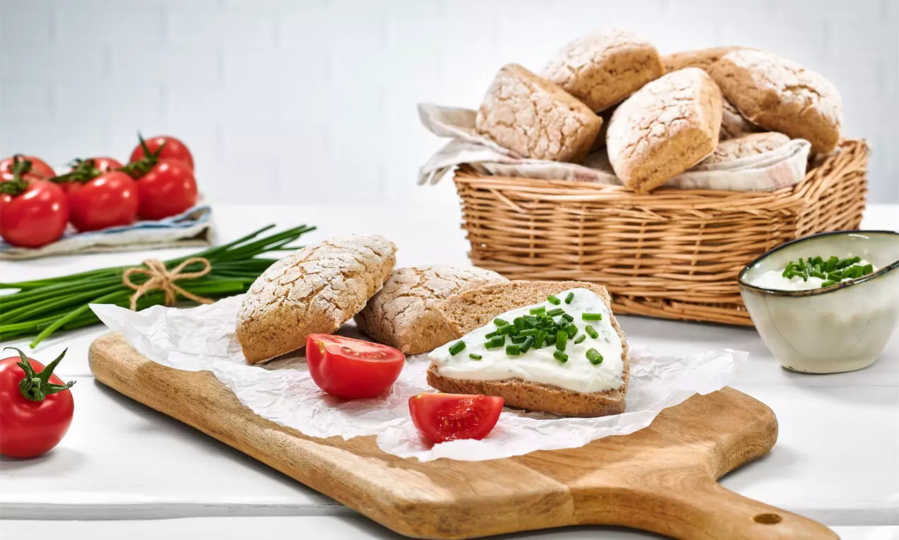 Roggenbrötchen mit mehliger Kruste auf einer Holzplatte, eines mit Frischkäse und Schnittlauch bestrichen, dazu halbierte Tomaten