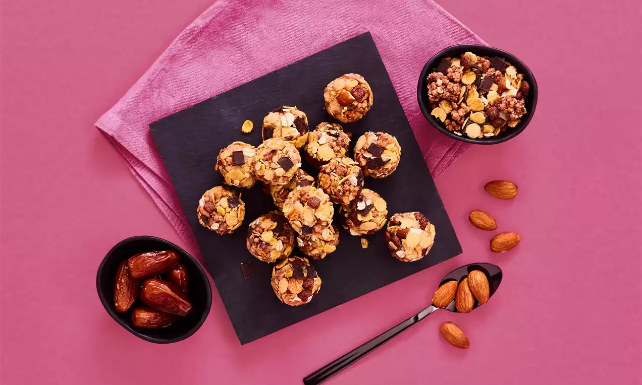 Protein Balls auf schwarzer quadratischer Platte, goldbraun mit gehackten Nüssen, daneben Datteln, Mandeln und Löffel auf pinkem Tuch