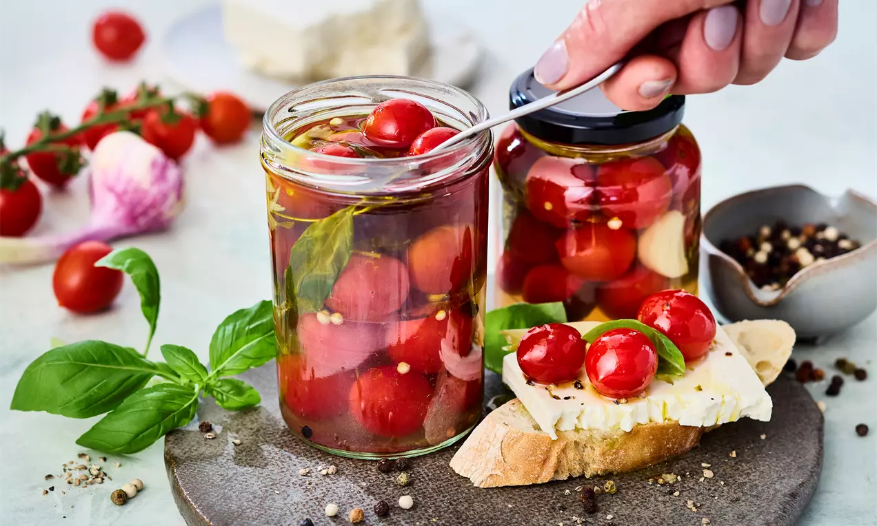 Eingelegte Tomaten in einem Glas mit frischen Kräutern, Knoblauchzehen und goldenem Olivenöl, leuchtend rot und saftig
