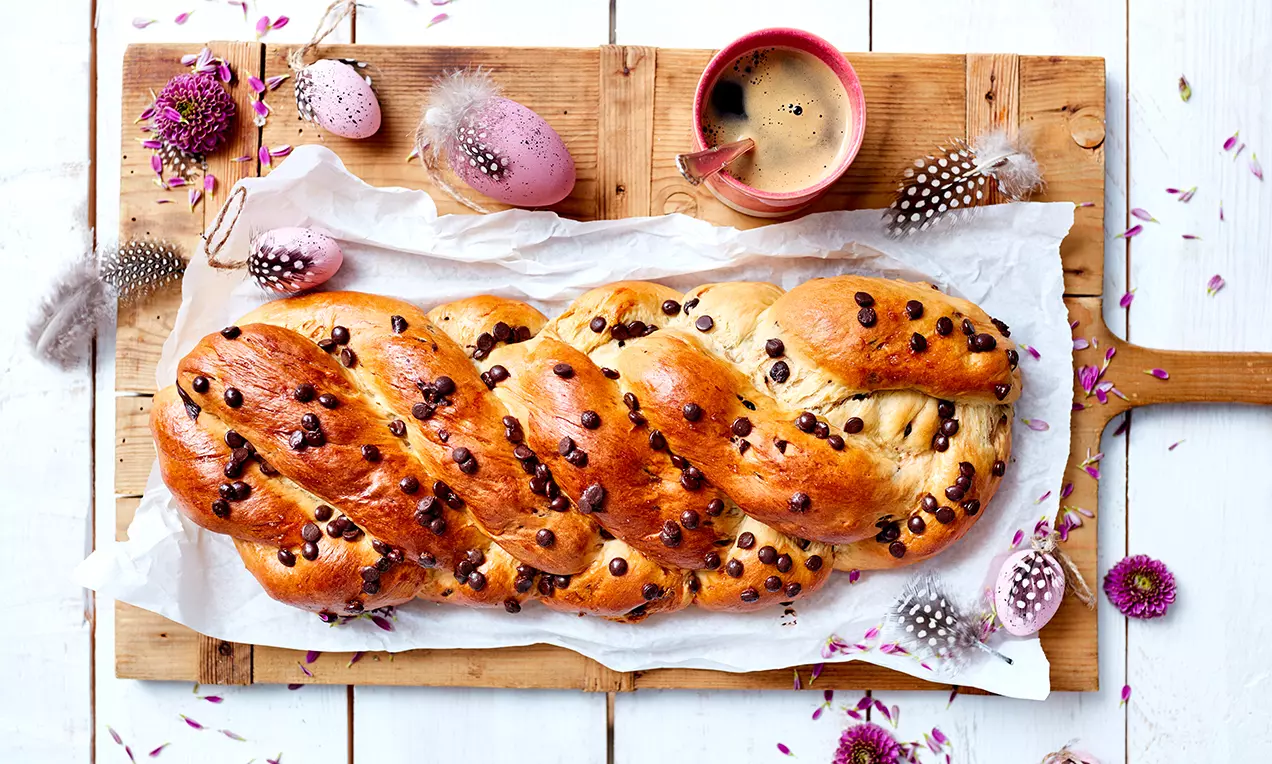 Glänzend goldbrauner Osterzopf aus 5 Strängen mit Schokotropfen auf Backpapier gelegt, dekoriert mit Federn, Eiern und Blumen auf Holzbrett