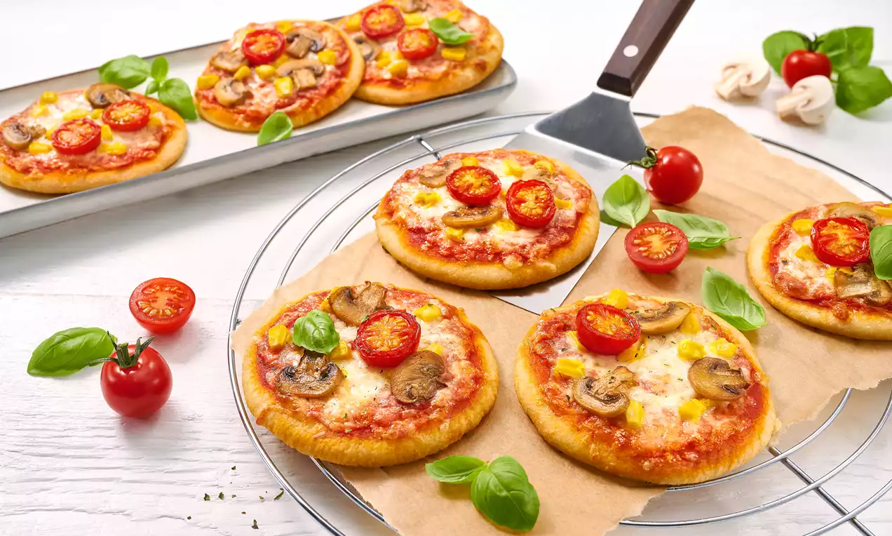 Kleine runde Pizzen mit roten Tomaten, braunen Champignons, gelbem Mais und grünem Basilikum auf Backpapier und Drahtgitter gelegt