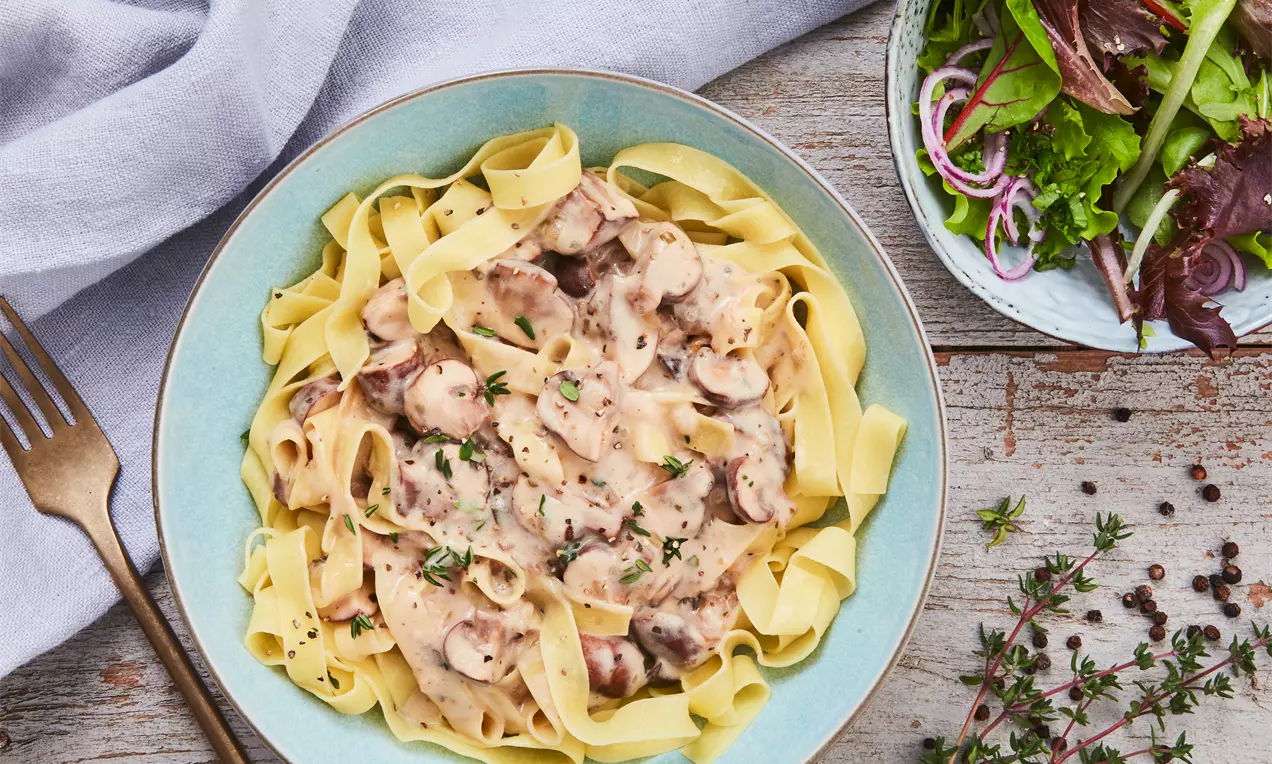 Nudeln mit Champignonsoße in weißer Schüssel, goldene Pasta mit cremiger Pilzsoße, grüner Petersilie und Käse garniert