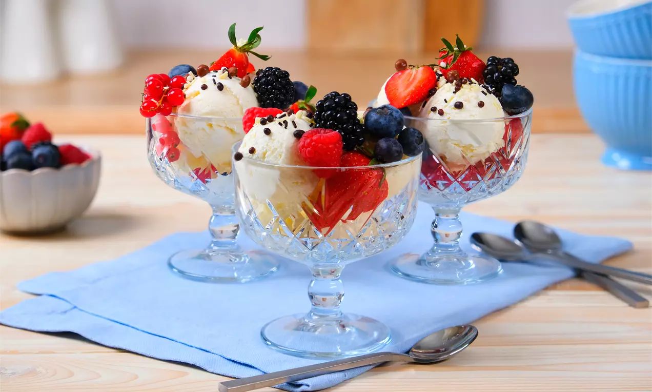 Frozen-Joghurt-Kugeln in kristallklaren Dessertgläsern, mit frischen Beeren und Schokostreuseln auf hellblauer Stoffserviette arrangiert