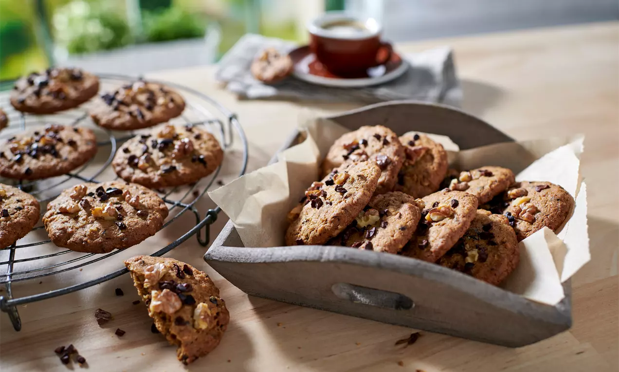 Dinkel-Cookies mit ungleichmäßig runder Form, goldbrauner Oberfläche, Walnussstücken und Schokolade auf Holzbrett und Gitter arrangiert
