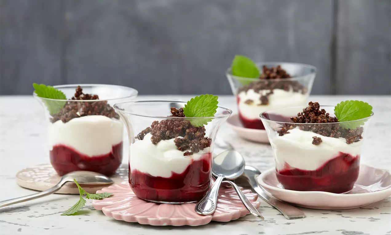 Gläser mit rotem Kompott, weißer Creme, knusprigen Schokobröseln und grünen Minzblättern auf rosa Tellern und heller Oberfläche