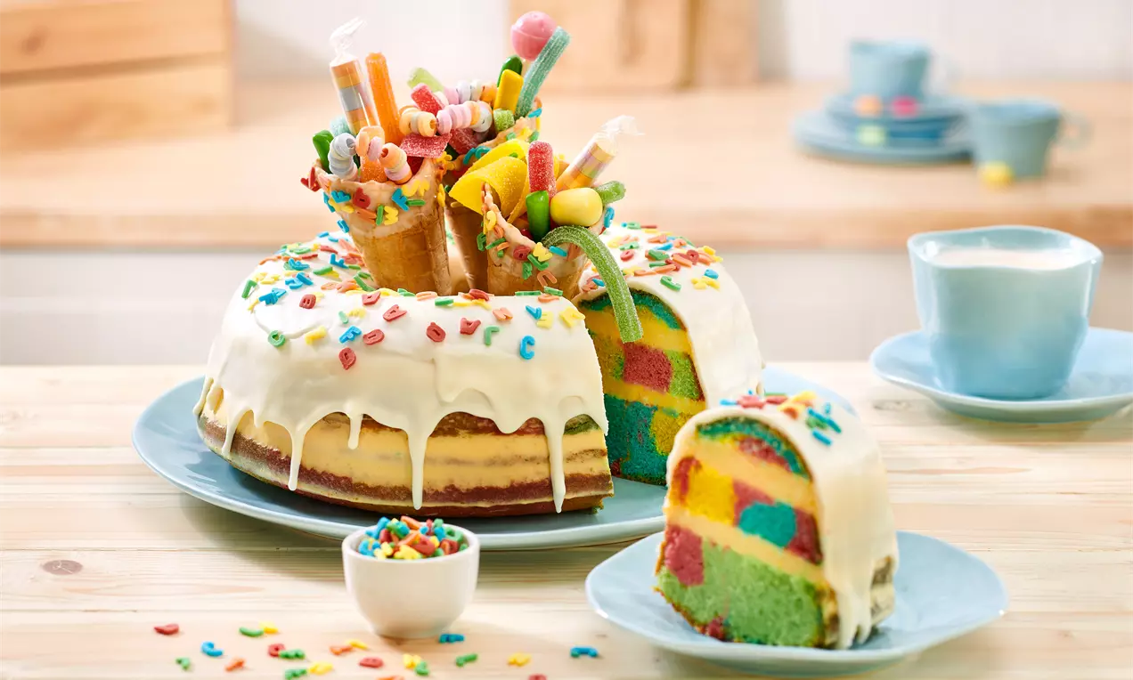 Bunte Zuckertüten-Torte mit weißer Glasur, farbigem Zuckerstreusel und süßen Naschereien, angeschnitten auf einem hellblauen Teller