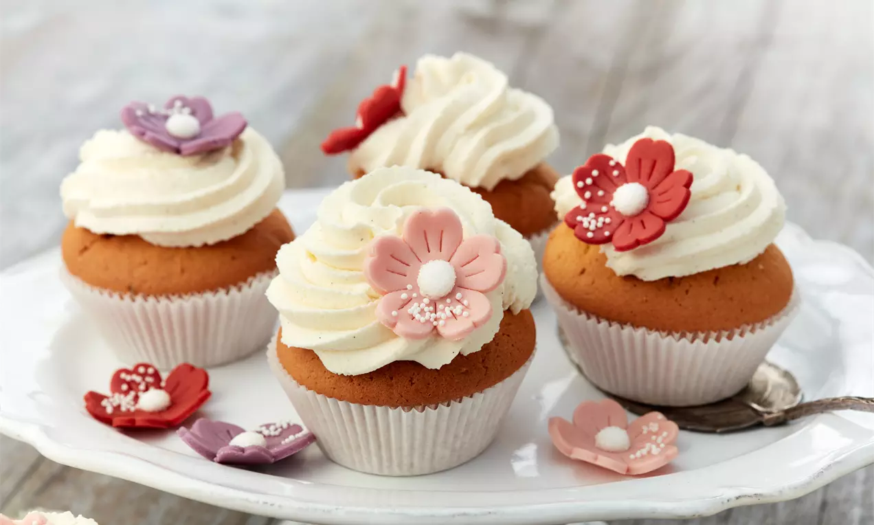 Vanille-Cupcakes mit geschwungener weißer Cremehaube, verziert mit bunten Zuckerblumen, angerichtet auf einer weißen Tortenplatte mit Löffel