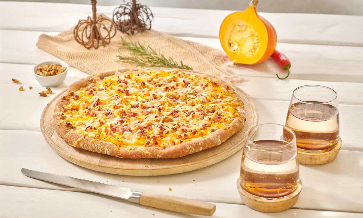 Goldbraune Pizza mit knusprigem Rand, belegt mit orangefarbenem Kürbis, Käse und Speckwürfeln auf rundem Holzbrett mit herbstlicher Dekoration