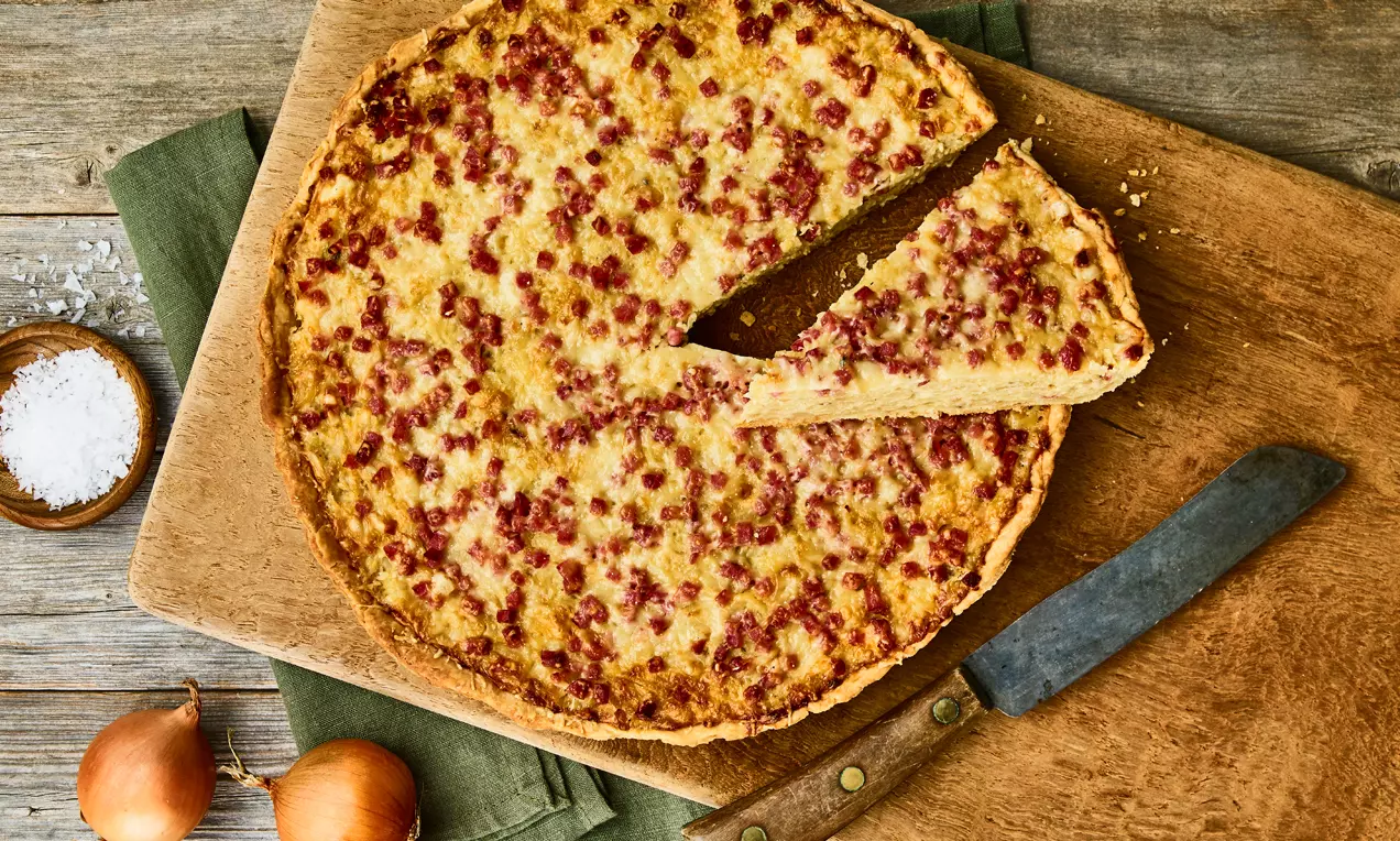 Runde goldbraune Wähe mit roten Speckstückchen, ein herausgeschnittenes Stück, angerichtet auf Holzbrett mit Messer, Zwiebeln und Salzschale