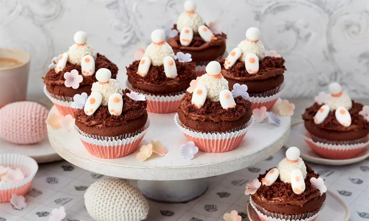 Schokoladenmuffins mit Osterhasen-Dekoration aus weißer Creme und Zuckerelementen, auf einem weißen Teller mit pastellfarbenen Blüten