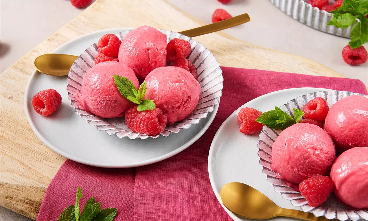 Kugeln aus intensiv rosafarbenem Himbeereis mit glatter Textur, angerichtet auf weißen Tellern mit frischen Himbeeren und Minzblättern