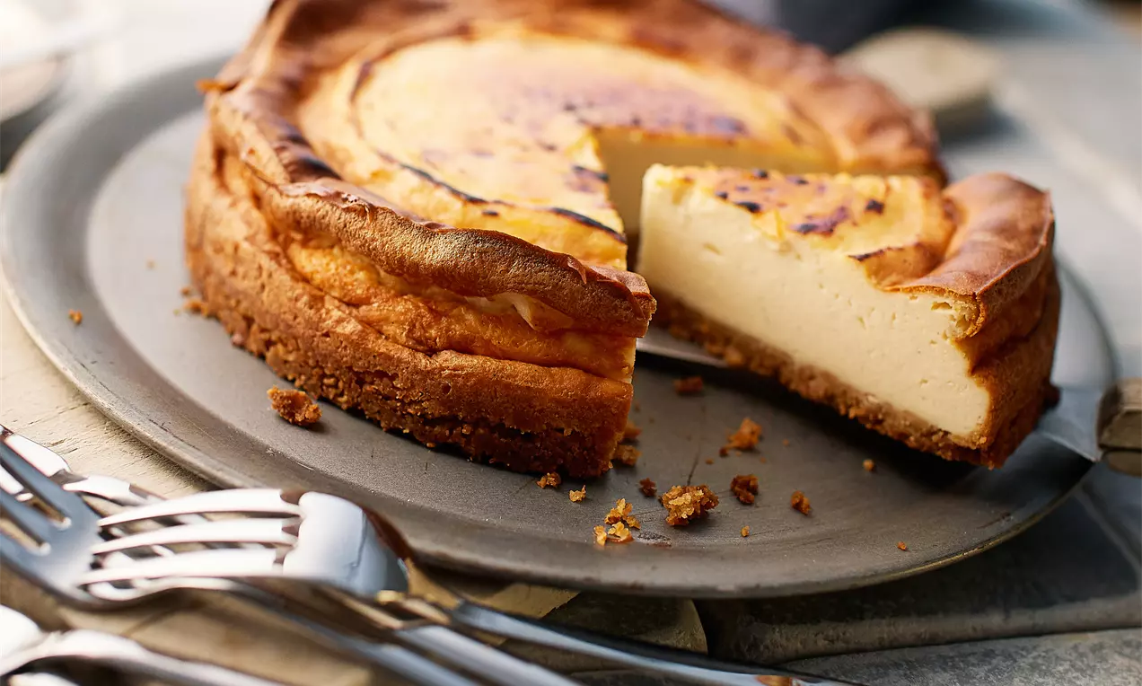Goldbrauner Cheesecake mit karamellisierter Oberfläche auf grauem Teller, angeschnittener saftiger Kern mit Krümeln und Besteck daneben