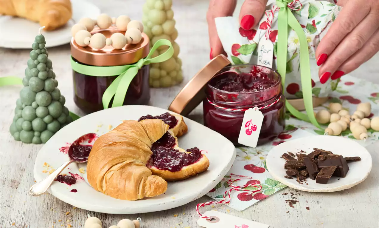 Kirschrote Marmelade mit cremiger Textur in einem offenen Glas, dekoriert mit grünem Band neben Croissant und Stückchen dunkler Schokolade
