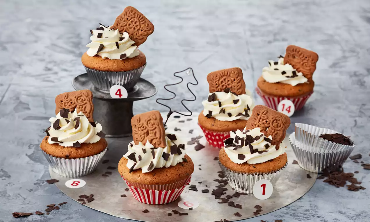 Spekulatius-Cupcakes mit goldbraunem Teig, dekoriert mit weißer Sahnehaube, Schokosplittern und einem Spekulatiuskeks auf silbernen und gemusterten Papierförmchen