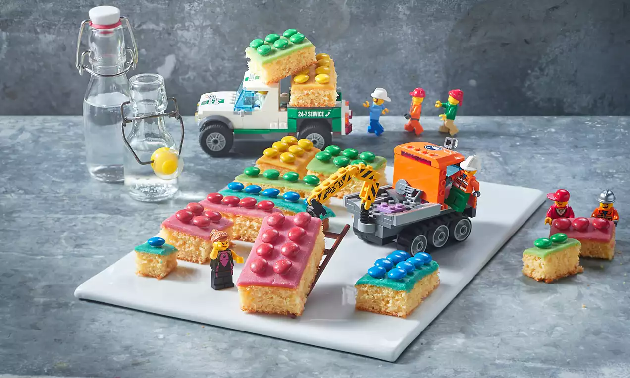 Bunte Kuchenstücke mit schokoladenüberzogenen Noppen, rechteckig geschnitten, auf weißer Platte, von Lego-Figuren und Fahrzeugen umgeben