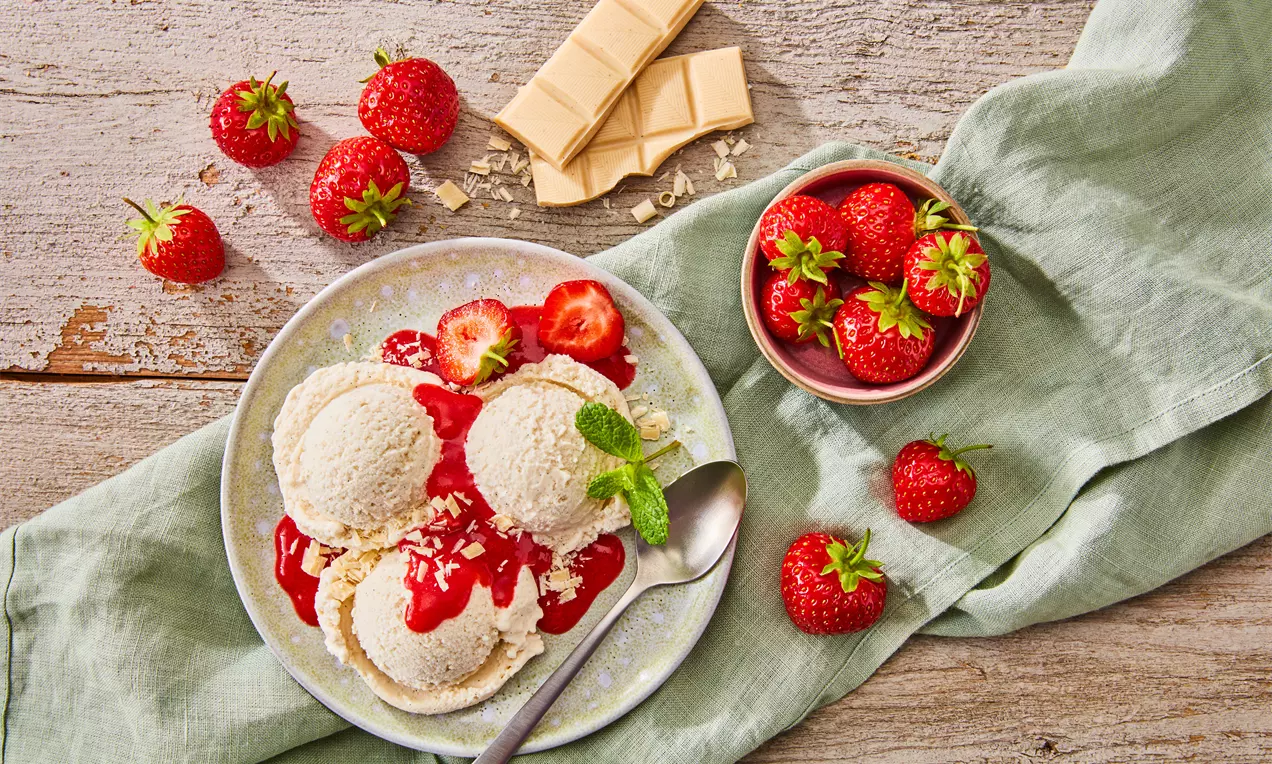 Drei Kugeln hellcremes veganes Vanilleeis mit glänzender, roter Erdbeersoße und Minzblatt auf hellem Teller, umgeben von frischen Erdbeeren