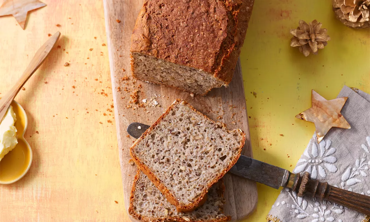 Mehrkornbrot mit goldbrauner Kruste und lockerer, körniger Textur, in Scheiben geschnitten auf einem rustikalen Holzbrett mit Besteck drapiert