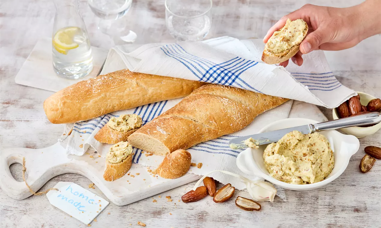 Cremiger, gelblich-bräunlicher Dip in einer weißen Schale neben frischem Baguette und halbierten Datteln auf einem Holzbrett mit Stofftuch