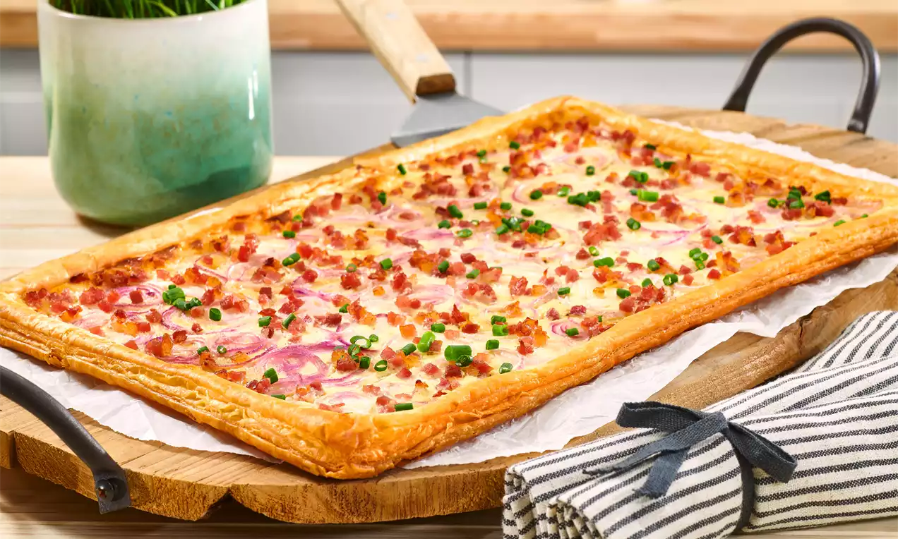 Rechteckiger Flammkuchen aus goldbraunem Blätterteig, belegt mit Speckwürfeln, roten Zwiebeln und grünen Frühlingszwiebeln auf Holzbrett