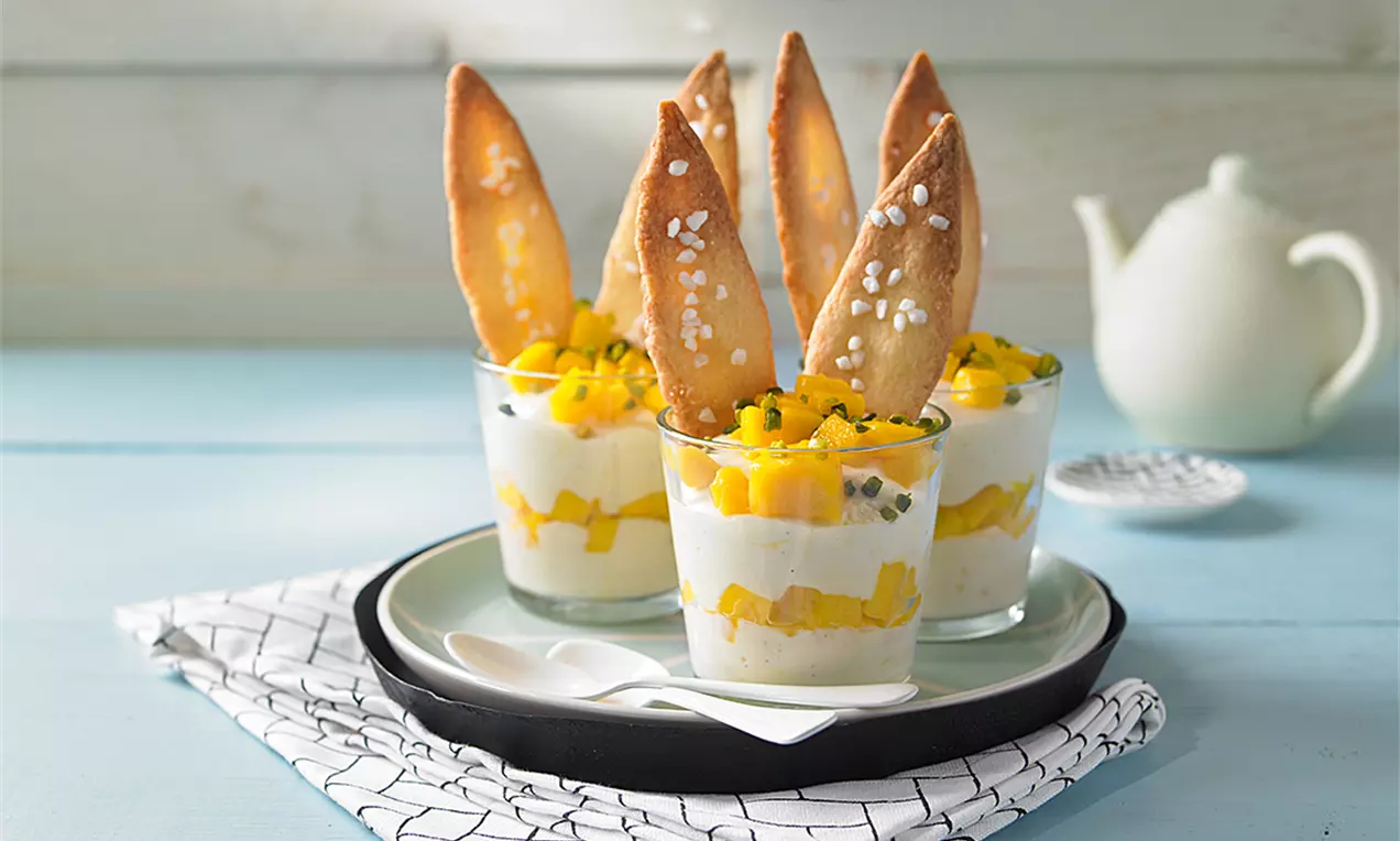 Mango-Mousse in Gläsern mit goldgelben Fruchtstücken, geschichtet auf weißer Creme, garniert mit knusprigen Keksstreifen und Sesamsamen