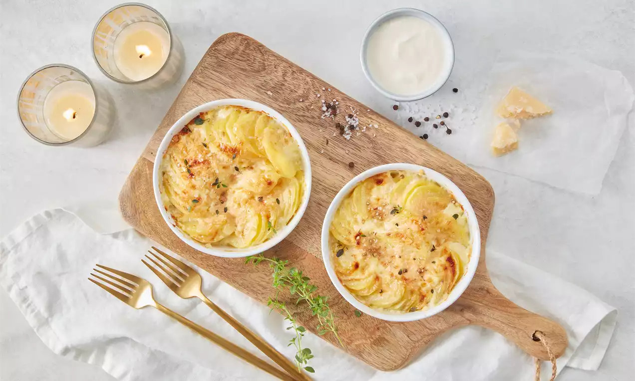 Goldbraunes Kartoffelgratin mit feiner Kruste, spiralförmig in weißen Schälchen arrangiert, auf Holzbrett mit goldenen Besteck und Thymian